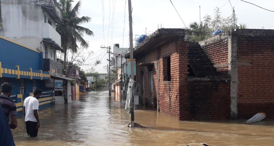 Zwei Männer stehen in einem Ort in Indien knietief im Wasser und schauen auf eine überflutete Straße