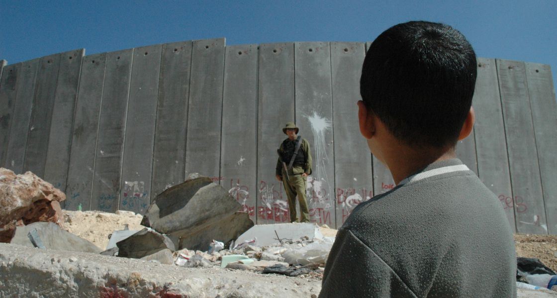 Auch dies hat der Bericht von Amnesty International deutlich gemacht: Israel und Palästina trennt viel mehr als eine hohe Mauer. Foto: Justin McIntosh (CC BY 2.0) Ein Junge steht vor einer riesigen Mauer, die von einem Soldaten beschützt wird.