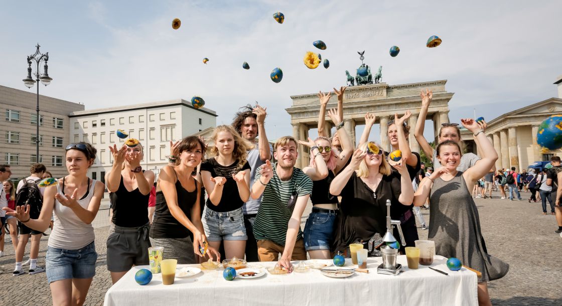 Ene Gruppe junger Menschen vor einem Tisch und im Hintergrund das Brandenburger Tor