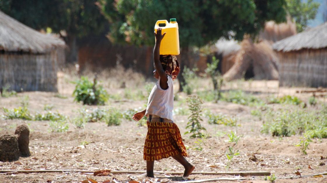 Ein Kind läuft durch ein Dorf und trägt einen Wasserkanister auf dem Kopf