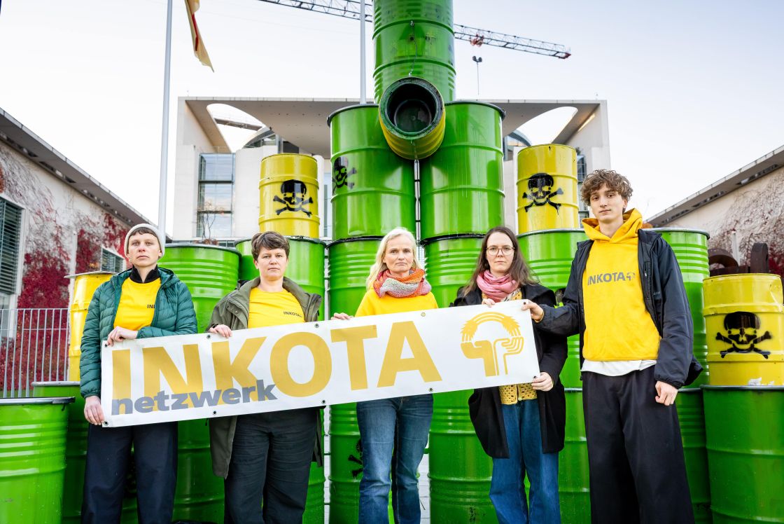 Fünf INKOTA-Aktivist*innen stehen vor gelben und grünen Fässern, sie halten ein INKOTA-Banner in der Hand