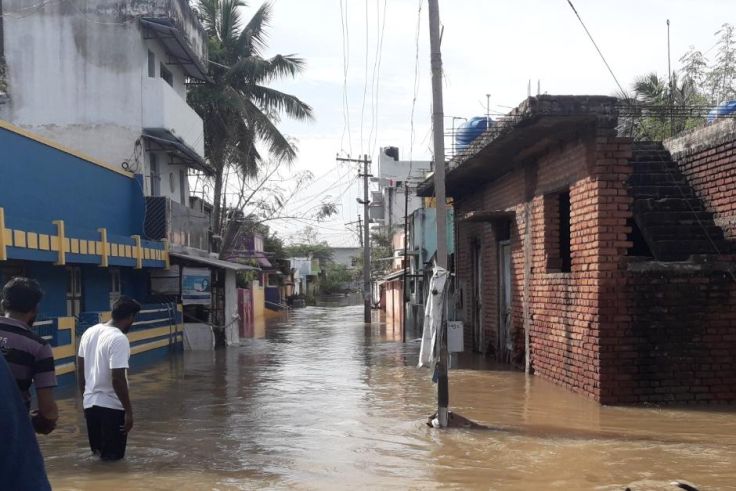 Zwei Männer stehen in einem Ort in Indien knietief im Wasser und schauen auf eine überflutete Straße