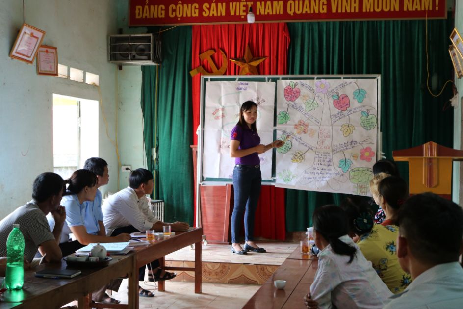 Eine Lehrerin vor einer Karte und merhere Erwachsene schauen sie an in einem Raum