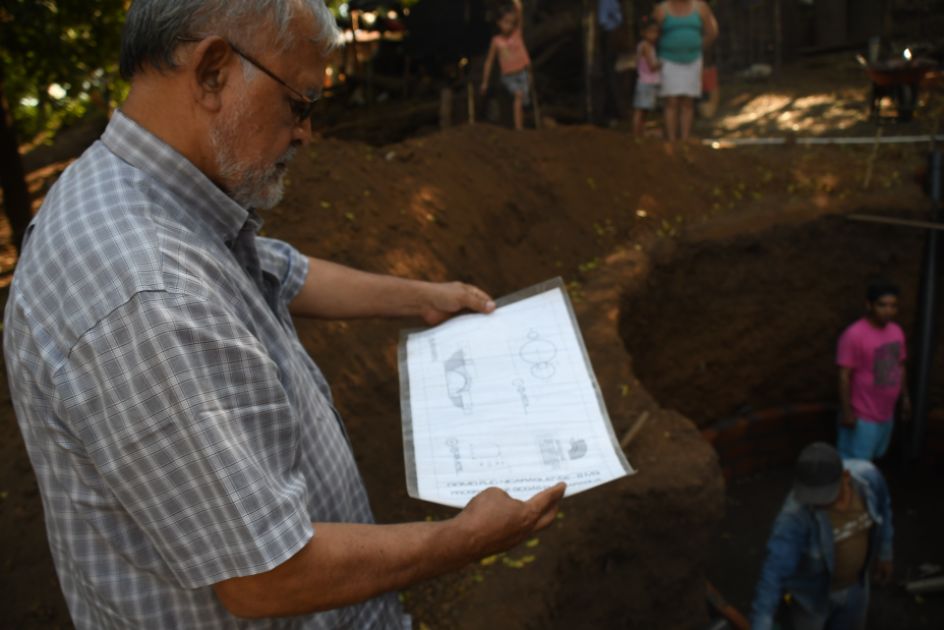 Ein Mann hält den Bauplan in den Händen und steht vor einer tief ausgehobenen Grube.
