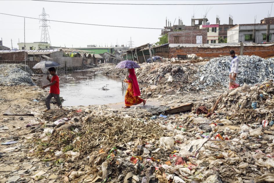 Zwei Personen mit Schirm passiren einen Fluss, an dessen Ufern haufenweise Müll liegt