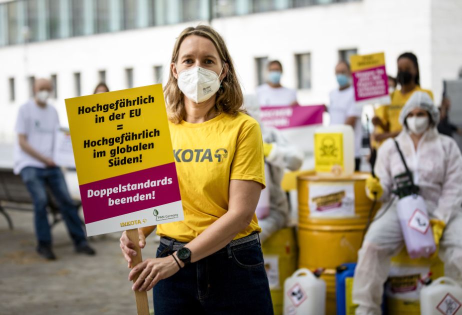 Eine Frau hält ein Schild mit der Aufschrift Hochgefährlich in der EU ist auch hochgefährlich im Globalen Süden. Dahinter mehrere Aktivistis mit Fässern und Kanistern, auf denen Totenköpfe und Warnhinweise geklebt sind.
