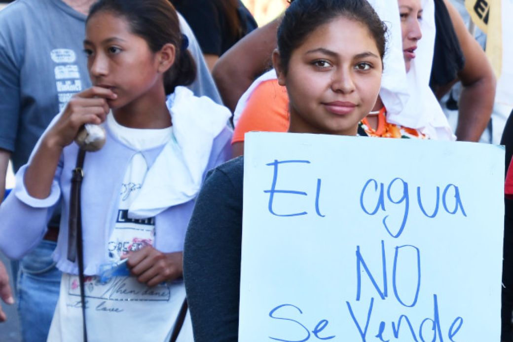 Zwei Frauen im Vordergrund einer Demo. Eine Frau hält ein Schild mit der Aufschrift: El agua no se vende - Das Wasser steht nicht zum Verkauf