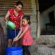 Eine Frau gießt aus einer Schüssel Wasser, daneben steht ein Kind und hält die Hände unter den Wasserstrahl, darunter eine blauer Tonne